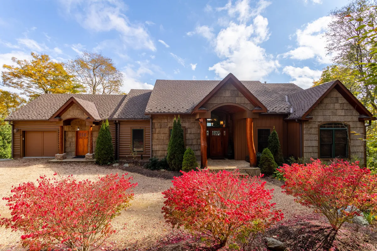 The image showcases the inviting entrance of Buckhorn Lodge, framed by vibrant red foliage in the foreground and a striking textured stone facade. The porch hints at relaxing moments spent taking in the fresh mountain air, while the spacious yard suggests gatherings and peaceful mornings in nature.