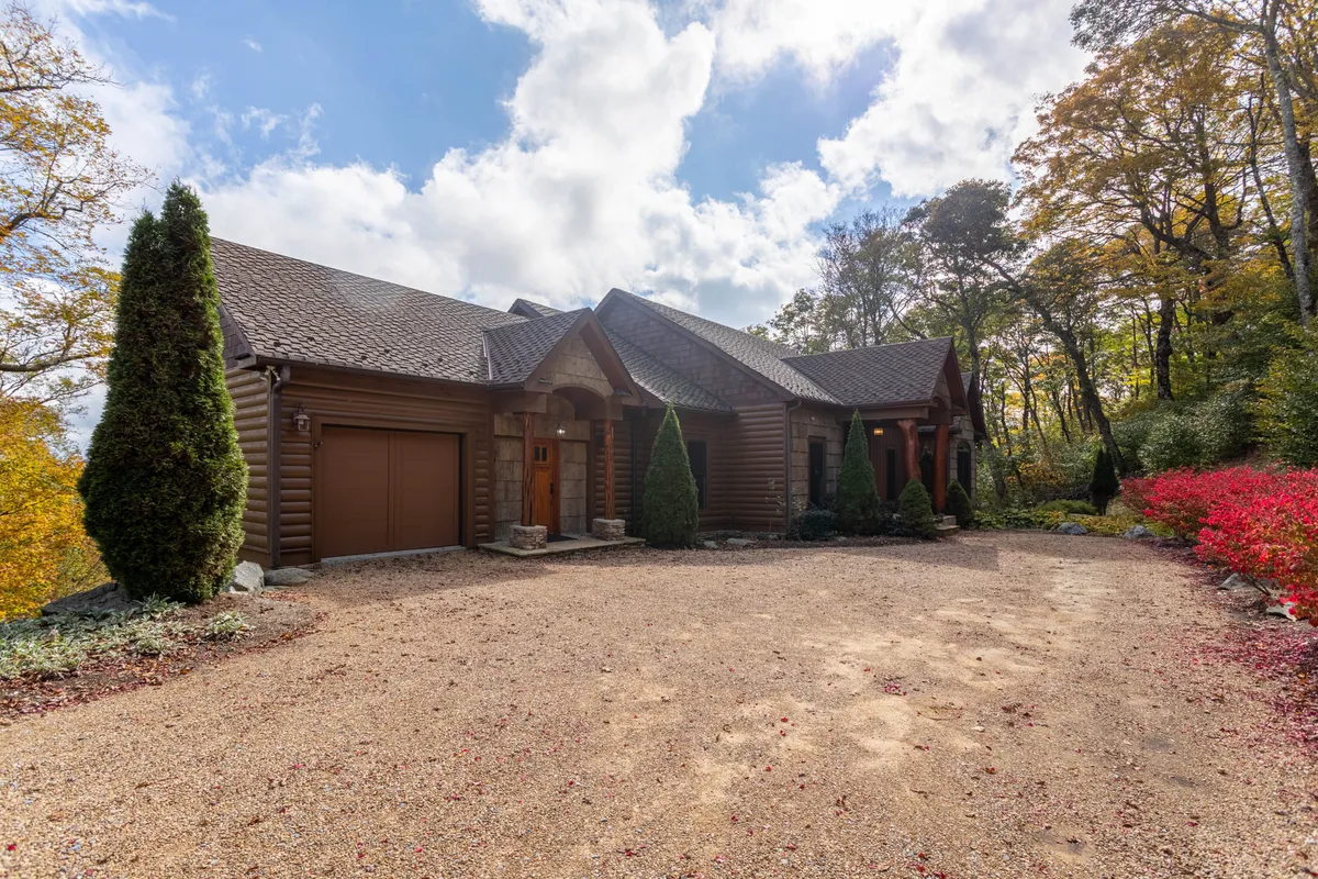 A charming log cabin structure set amidst a landscaped garden with colorful autumn foliage and a stone pathway. The porch offers spots for outdoor gatherings and quiet moments while watching the sunset over the mountains.