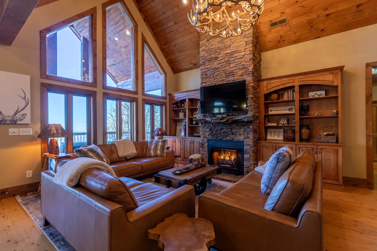 A spacious living room anchored by a large stone fireplace, with expansive windows framing panoramic mountain views. Leather sofas and a wooden chandelier create an inviting atmosphere, ideal for family gatherings and group activities like board games or movie nights.