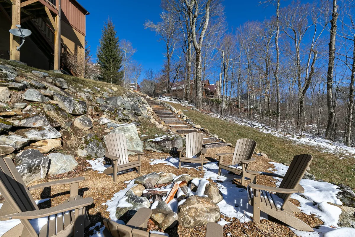 A cozy fire pit area with Adirondack chairs arranged on a stone pathway among trees, suggesting an inviting spot for evening gatherings, s'mores, and storytelling under the stars.