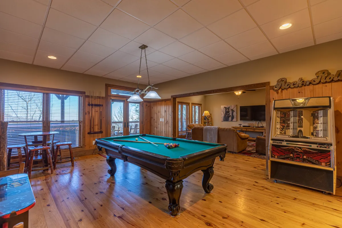 A lively game room with a central pool table set atop wooden floors, complemented by a bar area and large windows that fill the space with natural light. The informal, relaxed setup invites guests to socialize, play games, and enjoy drinks together. The overall ambiance is friendly and geared towards group entertainment.