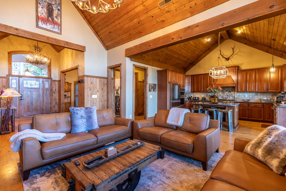 A spacious open living area featuring leather sofas gathered around a wooden coffee table, with views into the adjacent kitchen. A chandelier and rustic accents create a cozy atmosphere, while the arrangement of furniture supports group gatherings and shared activities.