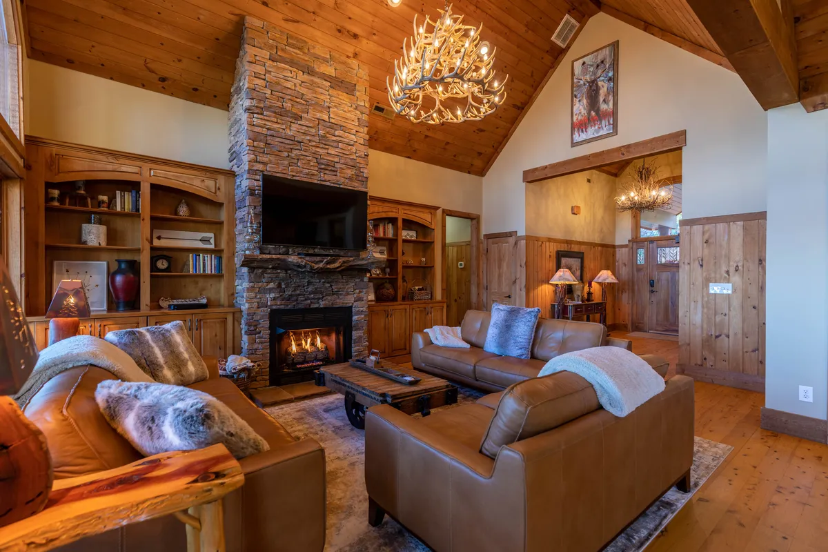 A warm, rustic interior featuring a stone fireplace, leather sofas, and wooden shelves. The space is arranged for group gatherings, with a chandelier adding a touch of elegance—ideal for movie nights or casual hangouts after a round of games.