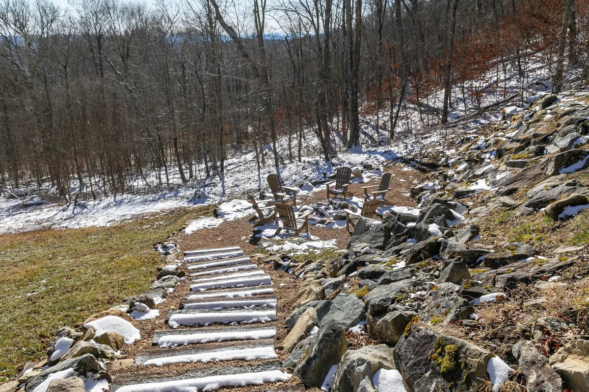 A tranquil outdoor seating area with Adirondack chairs circles a fire pit on a stone pathway, surrounded by trees and a gentle blanket of snow. The mood is peaceful, perfect for enjoying warm drinks or stargazing after a day on the slopes.