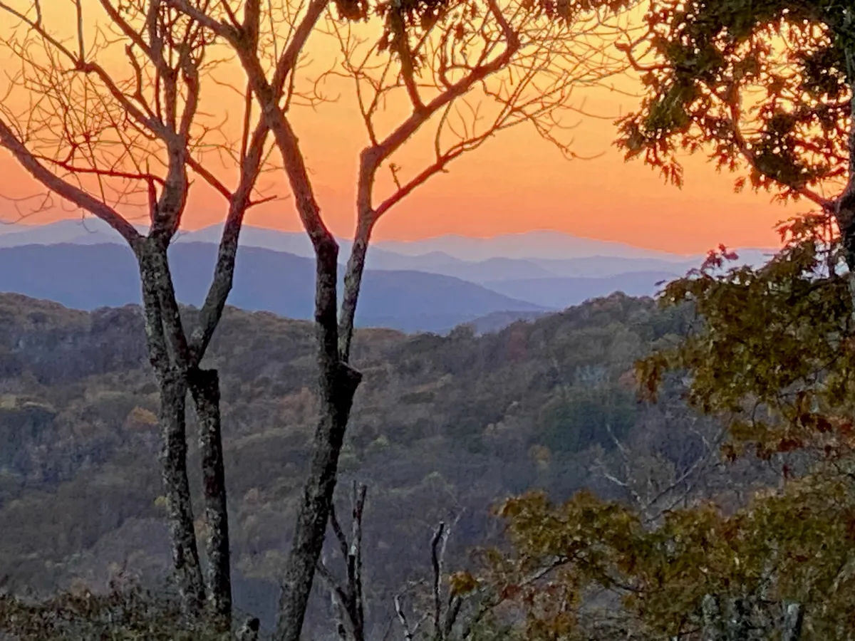 A sweeping sunset view with silhouetted trees in the foreground and distant Blue Ridge Mountains under a colorful sky. The tranquil scene evokes the vastness and natural beauty that guests enjoy while exploring the area. It captures the outdoor adventure and serenity promised by Buckhorn Lodge year-round.